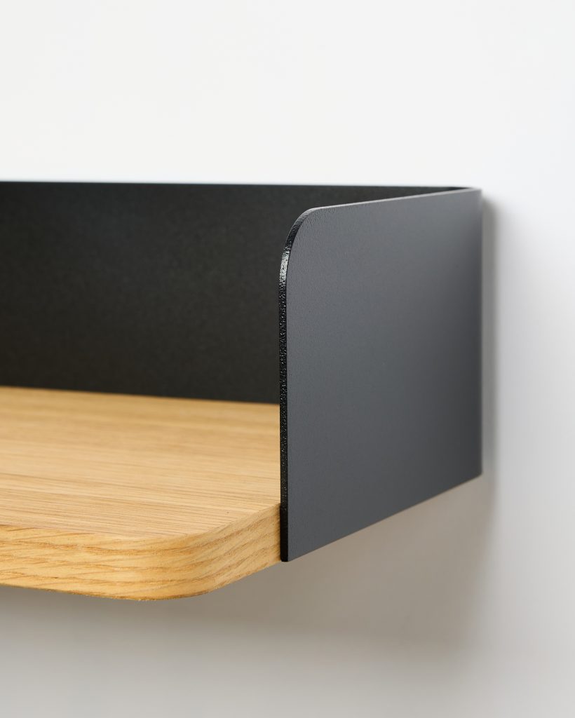WoodUpp wall shelf showing a natural oak shelf board with rounded edge and visible plywood grain, paired with a matte black powder-coated steel side and back rim forming a low guard and wall-mounted bracket—clean minimalist design emphasizing contrast between warm wood and dark metal.