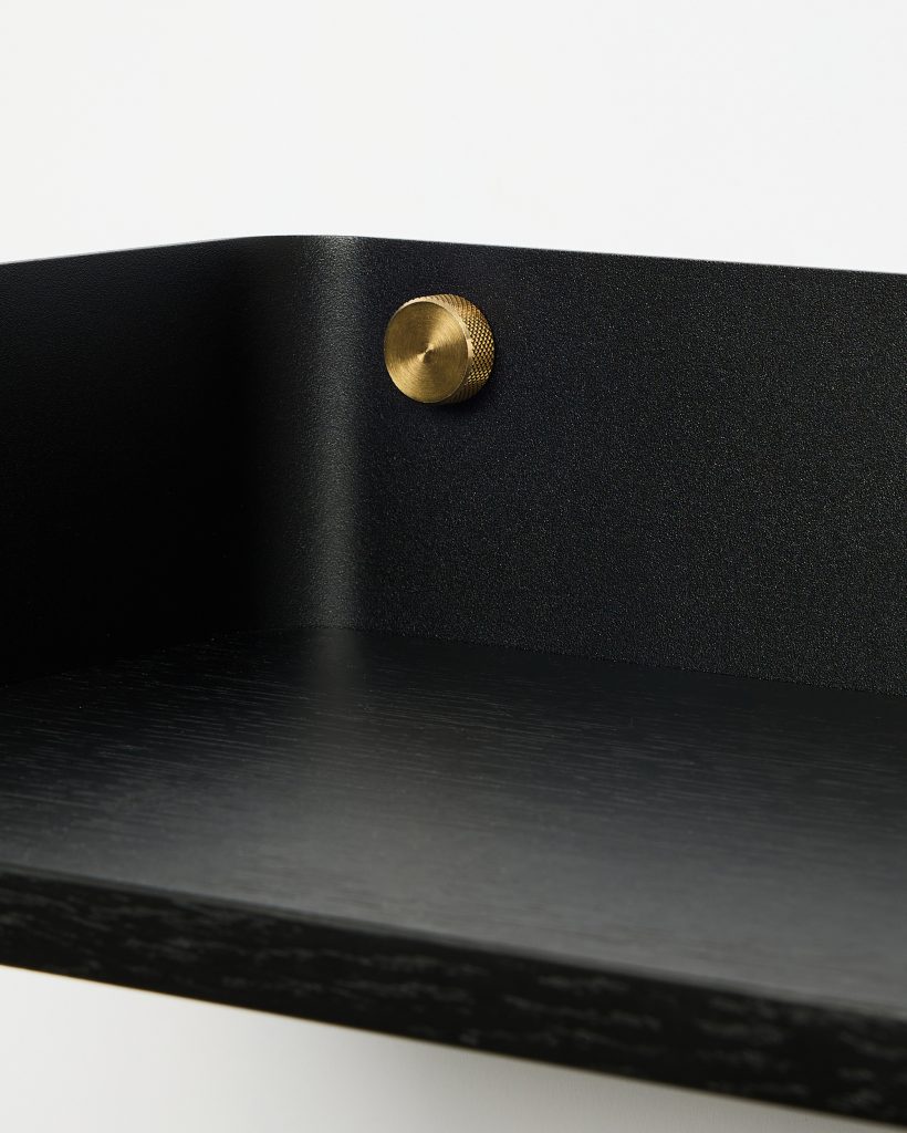 WoodUpp black wall shelf close-up showing a textured matte black metal backplate curving into a smooth black oak shelf, fastened by a single knurled brass screw. The minimalist WoodUpp shelf highlights wood grain on the platform and the contrast between dark timber and warm brass fittings.