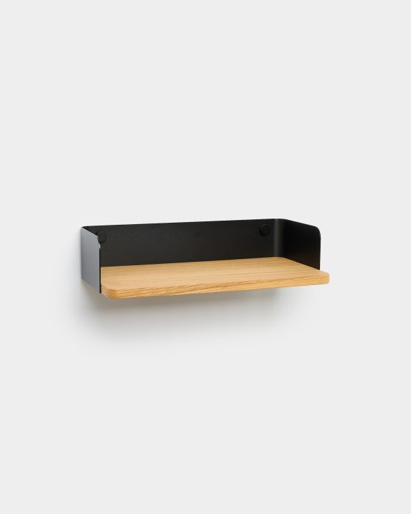 Minimalist WoodUpp wall shelf: a floating oak plank with visible grain and rounded front edge, seated in a matte black powder-coated metal tray back and sides with subtle round fixings, mounted on a white wall. Clean, modern shelf design suitable for small displays or storage.