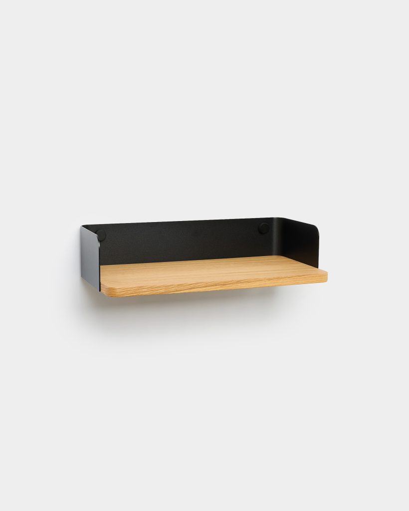 Minimalist WoodUpp wall shelf: a floating oak plank with visible grain and rounded front edge, seated in a matte black powder-coated metal tray back and sides with subtle round fixings, mounted on a white wall. Clean, modern shelf design suitable for small displays or storage.