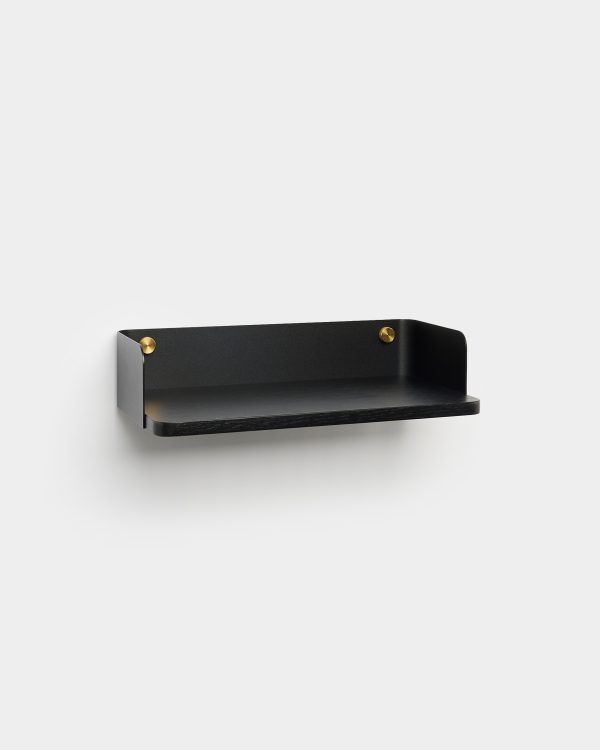 WoodUpp floating wall shelf in matte black: a slim rectangular black oak shelf with rounded front edge, backed by a curved matte-black metal panel and two polished brass mounting studs. Minimal, wall-mounted design for small objects, showing clean lines and subtle contrast between wood, metal and brass.