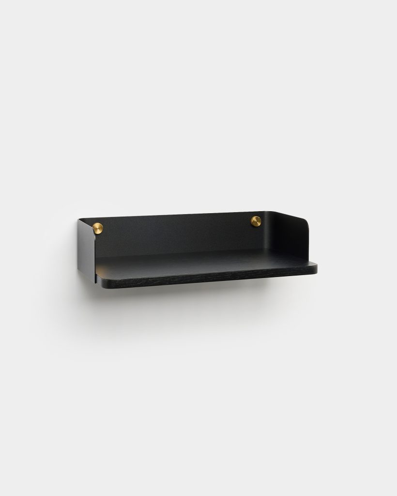 WoodUpp floating wall shelf in matte black: a slim rectangular black oak shelf with rounded front edge, backed by a curved matte-black metal panel and two polished brass mounting studs. Minimal, wall-mounted design for small objects, showing clean lines and subtle contrast between wood, metal and brass.