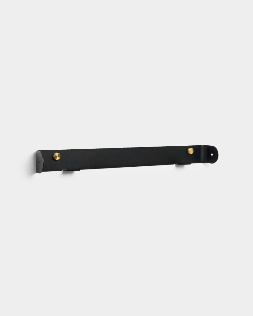 WoodUpp black metal wall rail mounted on a white wall, featuring two round brass fasteners and two slim fold-out hooks; minimalist, matte-finished horizontal strip designed for hanging coats, towels or accessories, with rounded end brackets and concealed screw holes for flush wall installation.