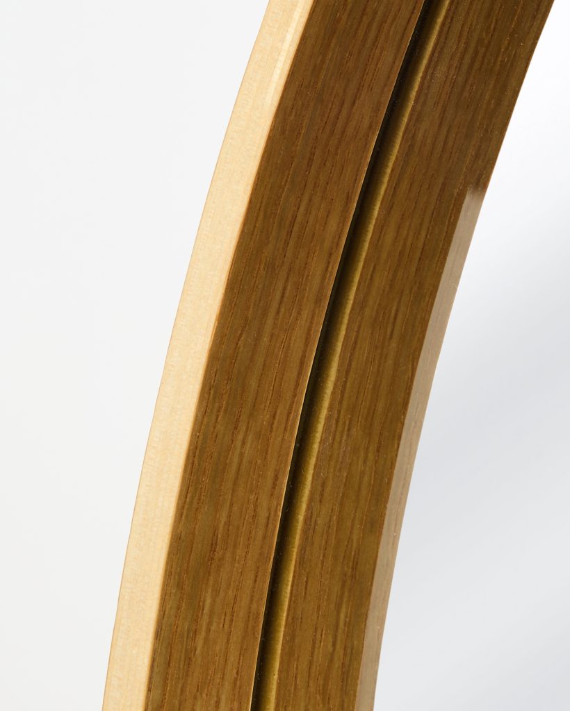 Close-up of a WoodUpp curved mirror frame showing layered light and warm oak wood veneers with a narrow brass‑colored inlay beside the reflective glass edge, highlighting smooth grain, rounded profile, and polished finish that showcases WoodUpp's premium craftsmanship and subtle depth.