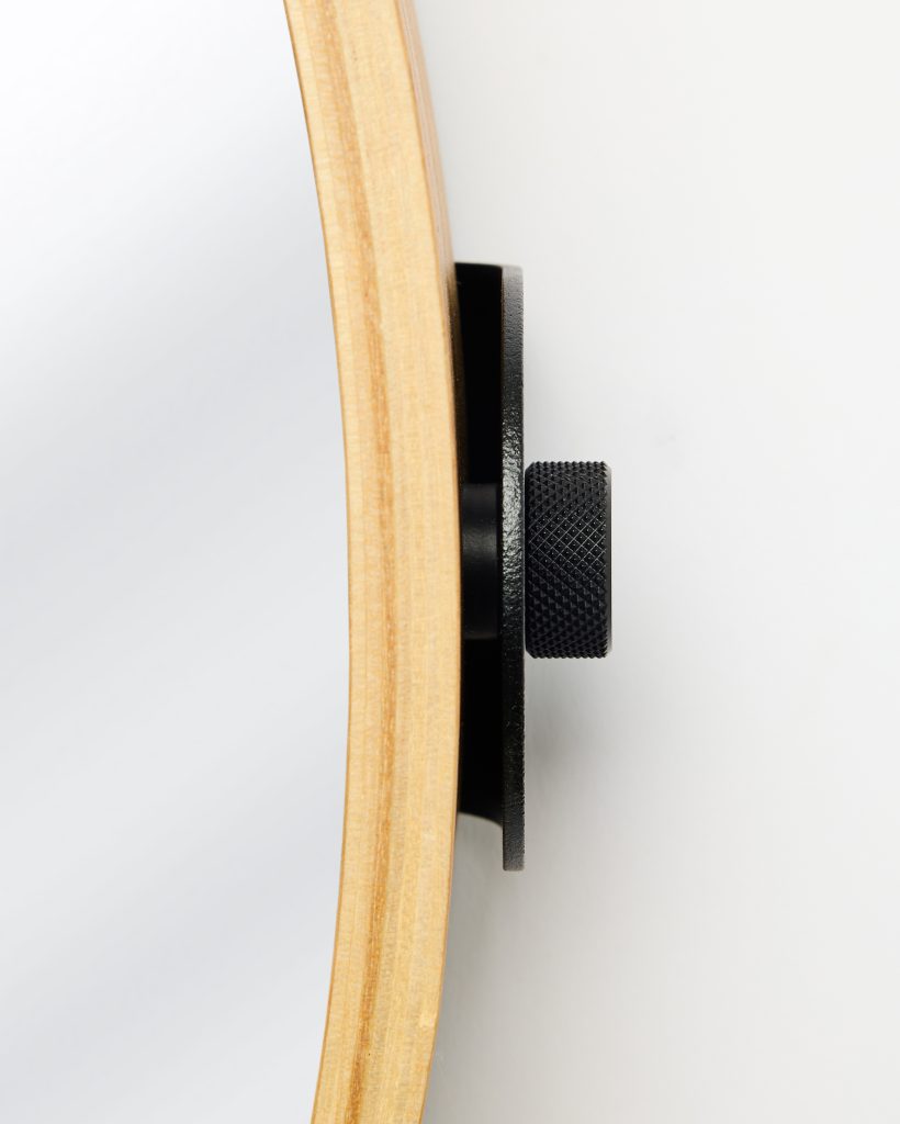 Close-up of a WoodUpp round wall mirror showing its light layered plywood frame and reflective glass edge, attached to a black powder-coated metal mounting plate with a textured knurled adjustment knob. The WoodUpp mirror and sturdy metal wall bracket are mounted flush against a white wall.