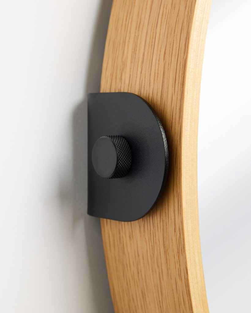 Close-up of a WoodUpp oak mirror frame showing a light oak veneer curved rim secured to the wall by a matte black WoodUpp metal wall bracket and knurled screw knob; visible wood grain, layered plywood edge and textured black finish emphasize material contrast and precise hardware detail.