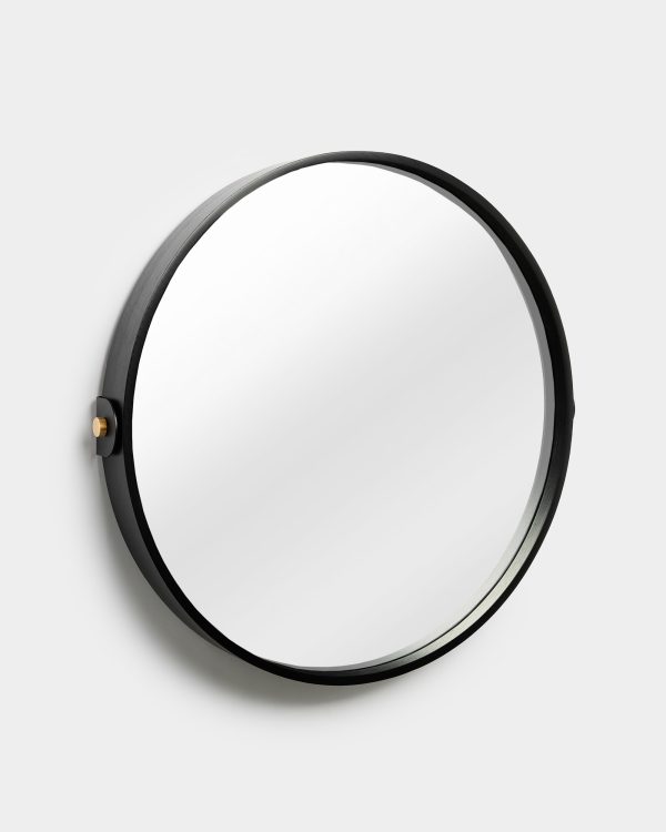 WoodUpp round wall mirror with a slim black-stained wooden frame, large circular reflective glass, and a small brass mounting peg on the left; photographed against a light grey wall, showcasing minimalist modern design, narrow profile and a soft, elegant shadow.