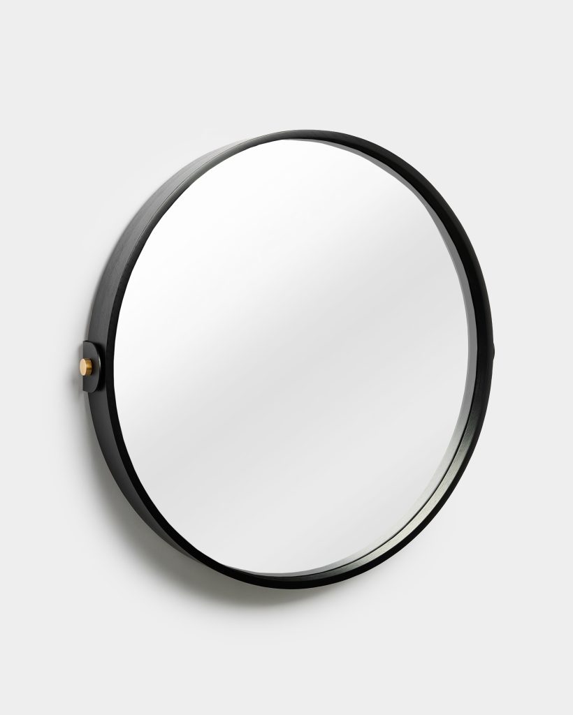 WoodUpp round wall mirror with a slim black-stained wooden frame, large circular reflective glass, and a small brass mounting peg on the left; photographed against a light grey wall, showcasing minimalist modern design, narrow profile and a soft, elegant shadow.