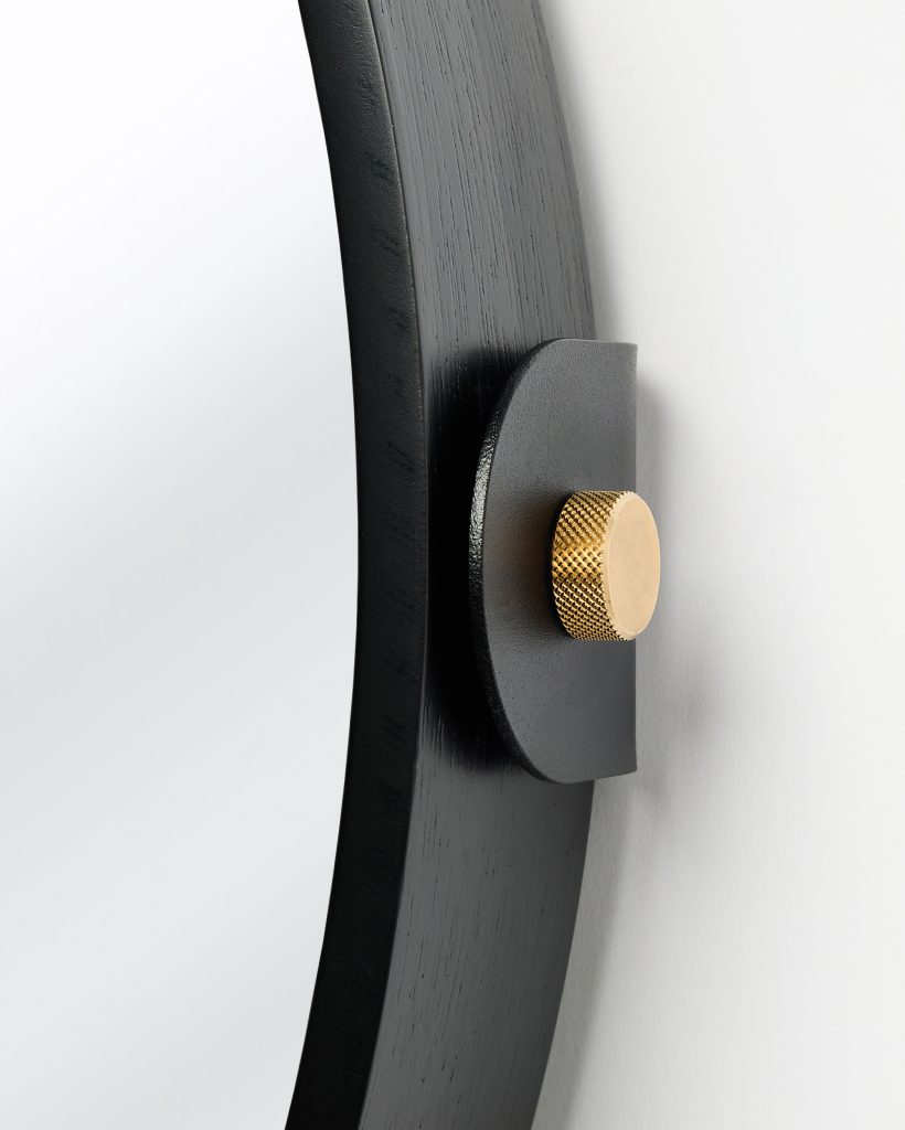 Close-up of a WoodUpp black oak mirror frame showing grain and matte finish, mounted by a black metal wall bracket and a textured knurled brass fastening knob. The image highlights WoodUpp’s round wooden mirror edge, minimalist metal bracket, and decorative brass mounting detail.
