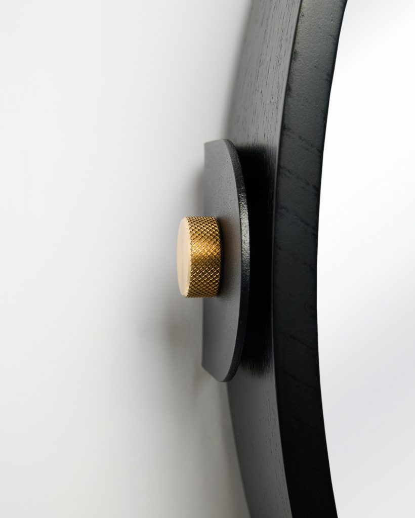 Close-up of a WoodUpp wall mirror edge showing a matte black wooden frame, curved mirror glass, a black metal mounting plate and a knurled brass-gold fixing knob. The WoodUpp mirror and its minimalist black bracket with textured golden fastener are highlighted against a plain white wall.