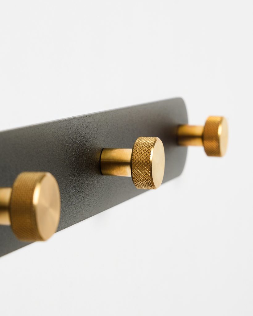 Close-up of a WoodUpp wall-mounted coat rack: a matte black rectangular metal backplate fitted with three cylindrical brass knobs. Each knob features a short stem and knurled textured circular head in warm gold finish, shown diagonally against a clean white background.