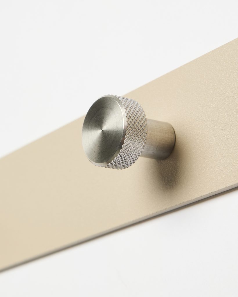 WoodUpp close-up of a stainless steel knurled wall peg mounted on a sleek matte beige WoodUpp metal strip; the round, brushed-steel knob features a textured grip and cylindrical stem, photographed at an angle against a white background highlighting hardware detail and finish.