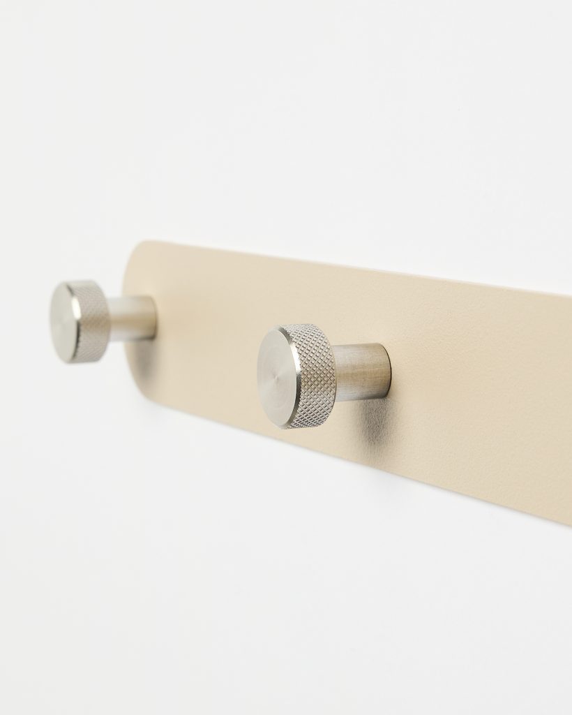 WoodUpp wall-mounted metal hook rail: a beige rectangular backplate with rounded ends fixed to a white wall, featuring two cylindrical stainless-steel pegs with knurled, disc-shaped ends. Minimal, modern coat or towel hooks from WoodUpp, shown in close-up, angled detail.