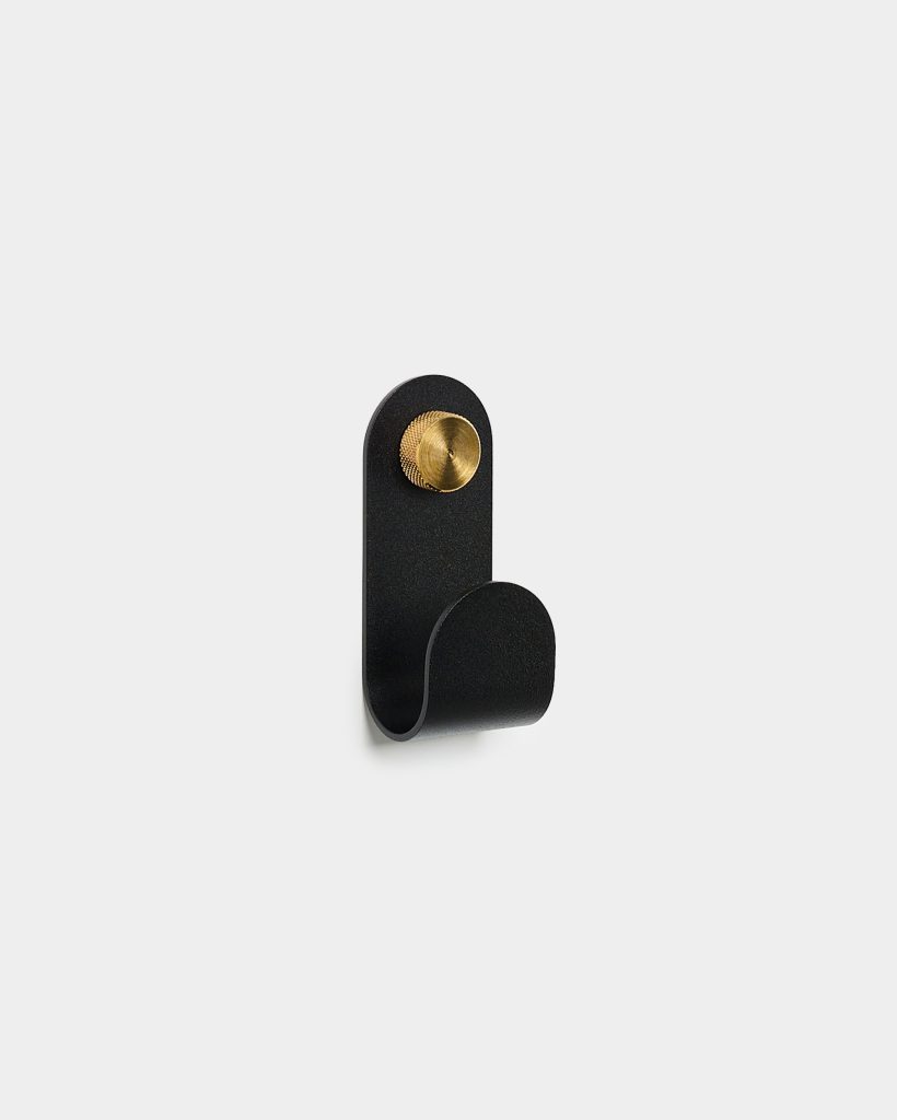 WoodUpp minimalist wall hook: a matte black, single-loop metal coat hook with rounded top, mounted by a knurled brass screw knob. The compact WoodUpp hook features a smooth curved profile for hanging coats, bags or towels, shown against a plain light background.