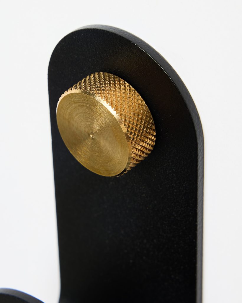 Close-up of a WoodUpp black metal wall hook featuring a rounded matte-black plate and a prominent knurled brass knob—circular, textured with diamond-cut pattern and concentric machining marks—used as a decorative fastening or hanging peg, highlighting contrasting finishes and tactile detail.
