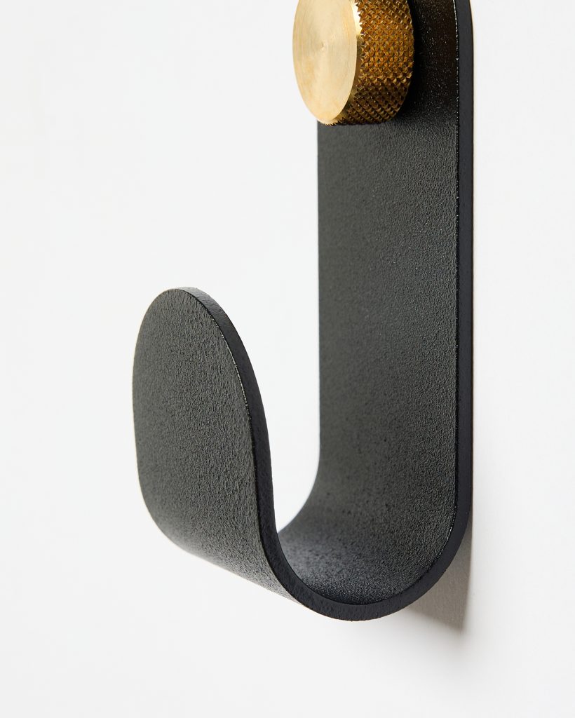 Close-up of a WoodUpp J-shaped wall hook: textured matte black steel curved hook mounted on a white wall, paired with a knurled brass WoodUpp mounting cap at the top. The minimalist hardware highlights clean lines, solid construction and tactile metal finishes.