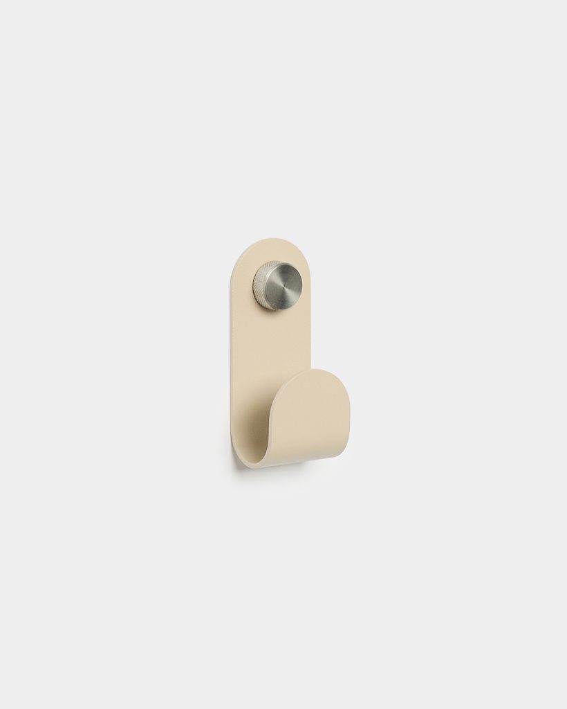 WoodUpp beige wall hook: a single minimalist metal hook with a rounded rectangular backplate and a curved front lip, matte sand-colored finish, mounted by a polished knurled stainless-steel screw. Simple Scandinavian design for hanging coats, towels, or bags, shown alone on a white background.