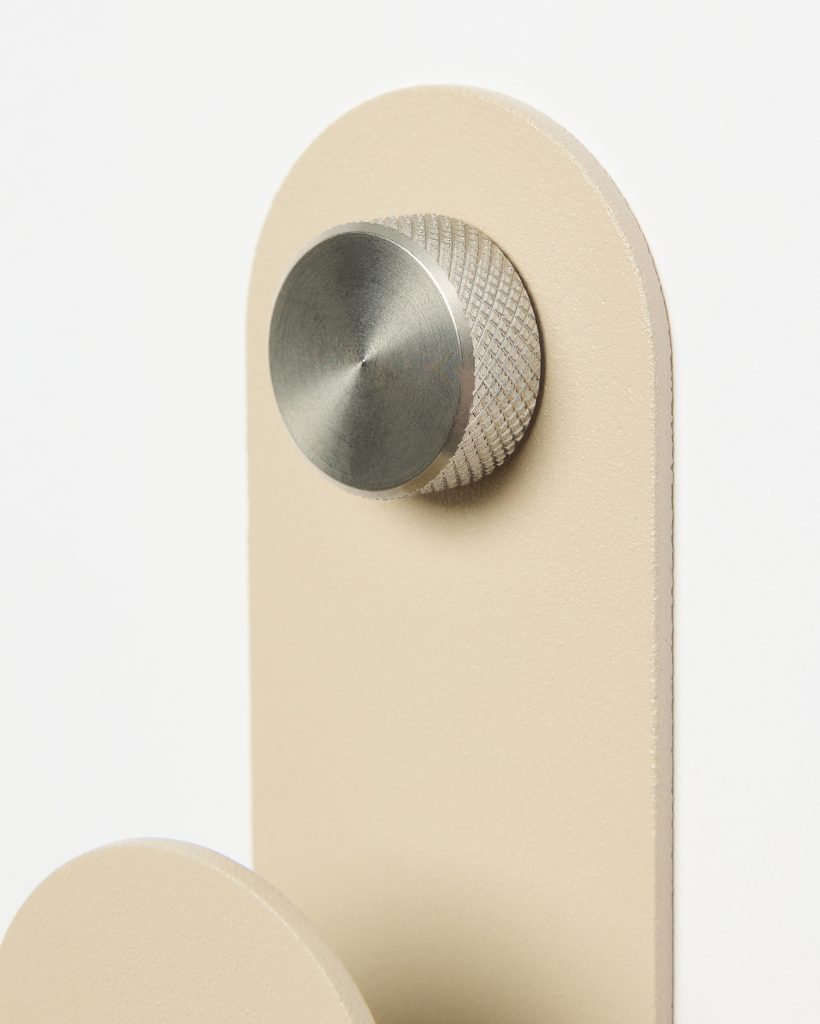 Close-up of WoodUpp wall hook: a matte beige, arched backplate featuring a knurled stainless‑steel cylindrical knob with a brushed circular face, and a lower matching beige circular peg partially visible. Minimal, modern finish highlights texture, precise metal detailing and subtle shadowing.