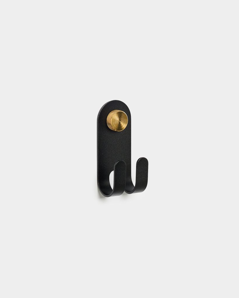 WoodUpp wall-mounted double hook: a matte black metal plate with two curved hooks and a rounded top, secured by a textured brass screw-cap, shown mounted on a white wall. Minimal, modern coat and accessory hanger from WoodUpp, compact, space-saving design.