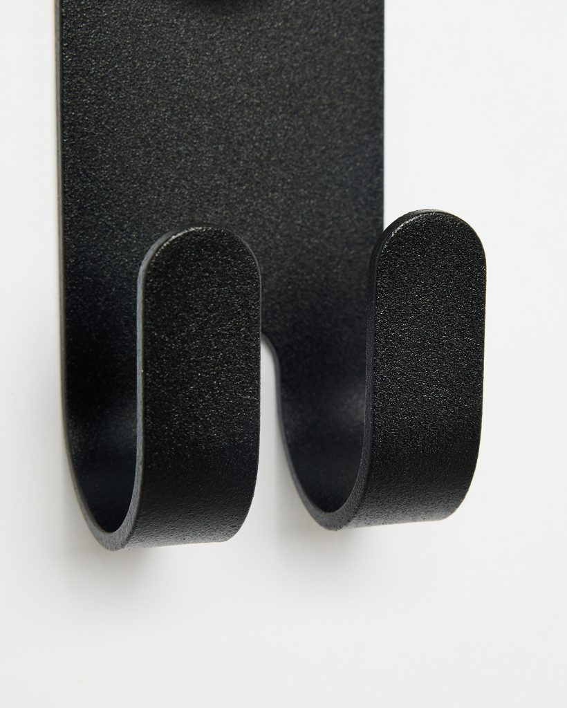 Close-up of a WoodUpp matte black double wall hook: a textured powder-coated steel backplate with two rounded U-shaped hooks protruding, forming twin downward-curving arms with smooth, rounded tips, mounted flush against a white wall—simple, sturdy design for hanging coats, bags, or towels.