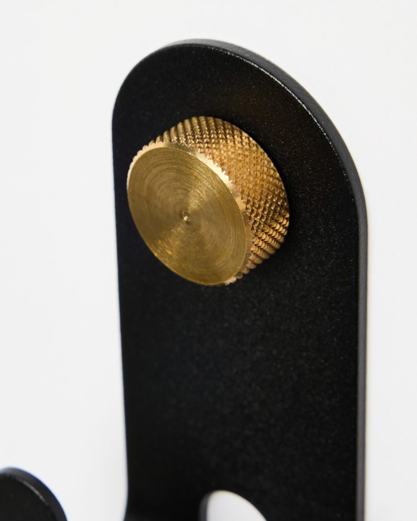 Close-up of a WoodUpp wall hook: matte black metal hanger with rounded top and integrated cutout, featuring a polished circular brass knurled knob protruding from the face. Clean white background highlights the contrasting black hook and warm brass accent and precise machining details are visible.