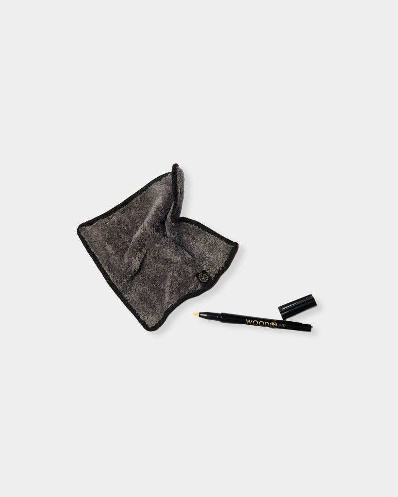 WoodUpp grey microfiber polishing cloth with black stitched trim and small round logo tag, showing plush texture, beside a glossy black WoodUpp-branded touch-up marker pen with gold lettering — cap removed to reveal a yellow felt tip — arranged on a plain white background.