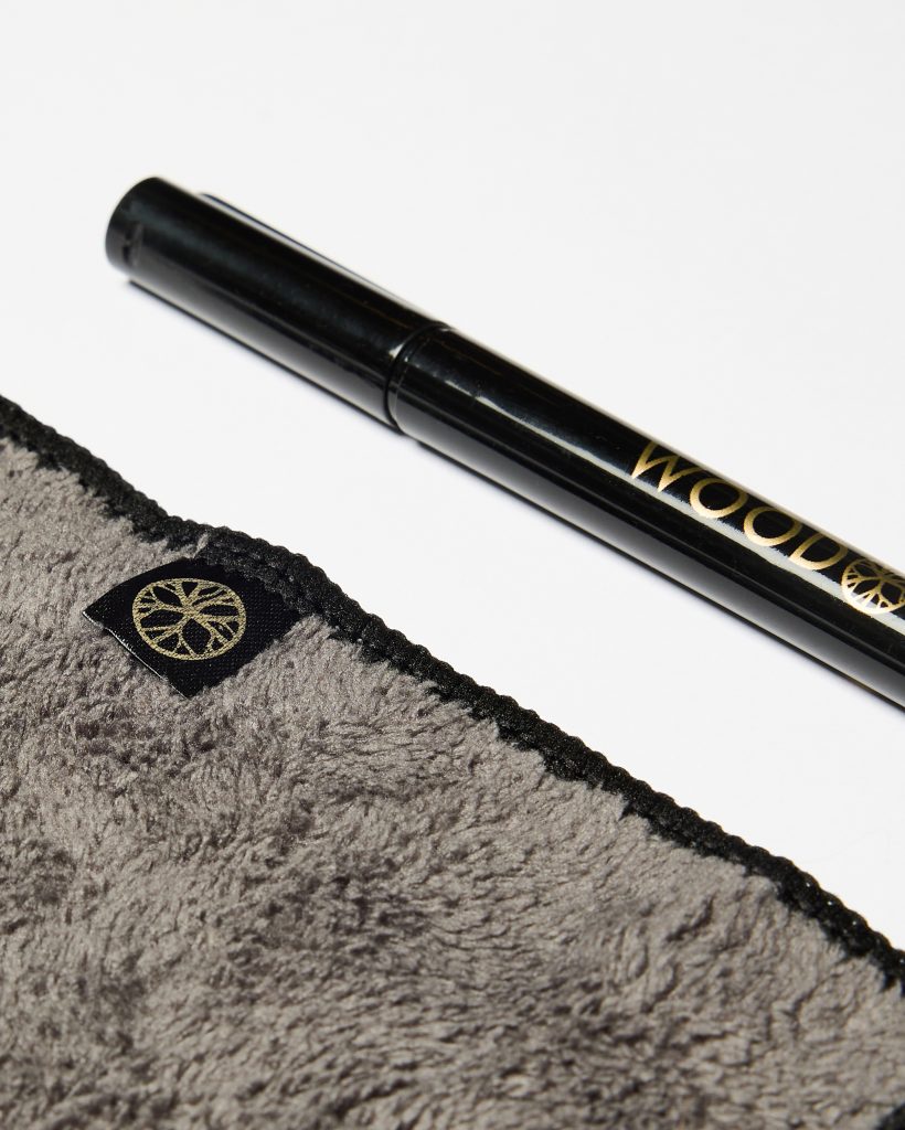 WoodUpp products on a white background: a glossy black WoodUpp detailing pen with gold branding positioned above a grey plush microfiber polishing cloth, featuring a black stitched edge and a small black tag printed with WoodUpp’s gold circular logo emblem.