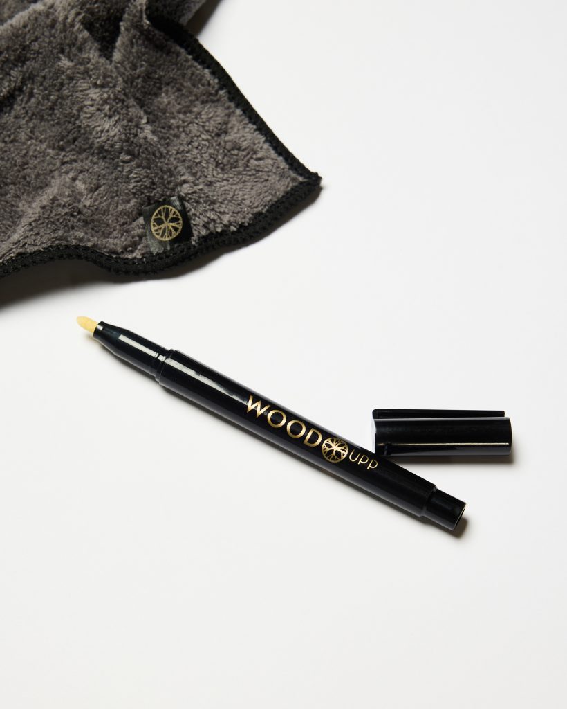 WoodUpp products: a sleek black WoodUpp touch-up pen with gold logo and a beige applicator tip, shown uncapped with cap beside it, alongside a soft dark gray microfiber polishing cloth with black stitched edge and a small WoodUpp logo tag.