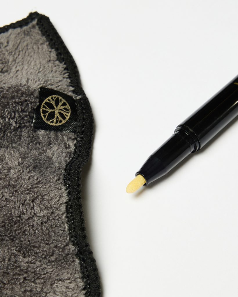 Close-up of WoodUpp products: a soft gray microfiber polishing cloth with black stitched edge and small black tag bearing a gold logo, beside a sleek black WoodUpp touch-up pen featuring a pointed yellow sponge applicator tip for applying finish or stain.