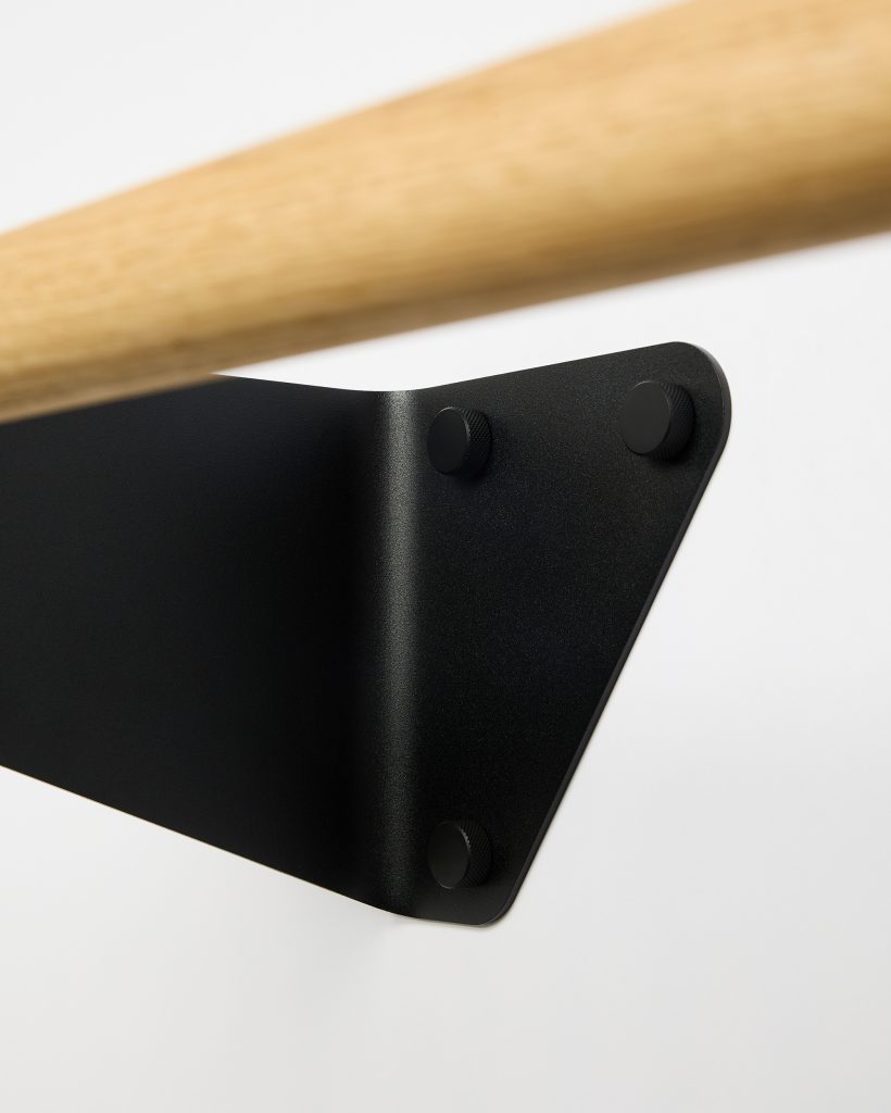 Close-up of a WoodUpp oak hanging rail paired with a matte black triangular wall bracket. The image shows the smooth light wood rail and textured powder-coated metal bracket with three knurled round black mounting caps, highlighting the brand's clean, minimal Scandinavian hardware design.