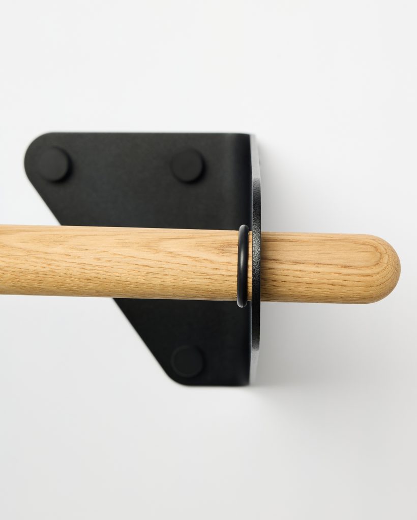 WoodUpp close-up of a natural oak dowel and matte black wall bracket: a smooth, rounded oak rod with visible grain threaded through a minimalist black powder-coated metal bracket, secured by a small black ring stop and three circular screw covers, shown against a clean white background.