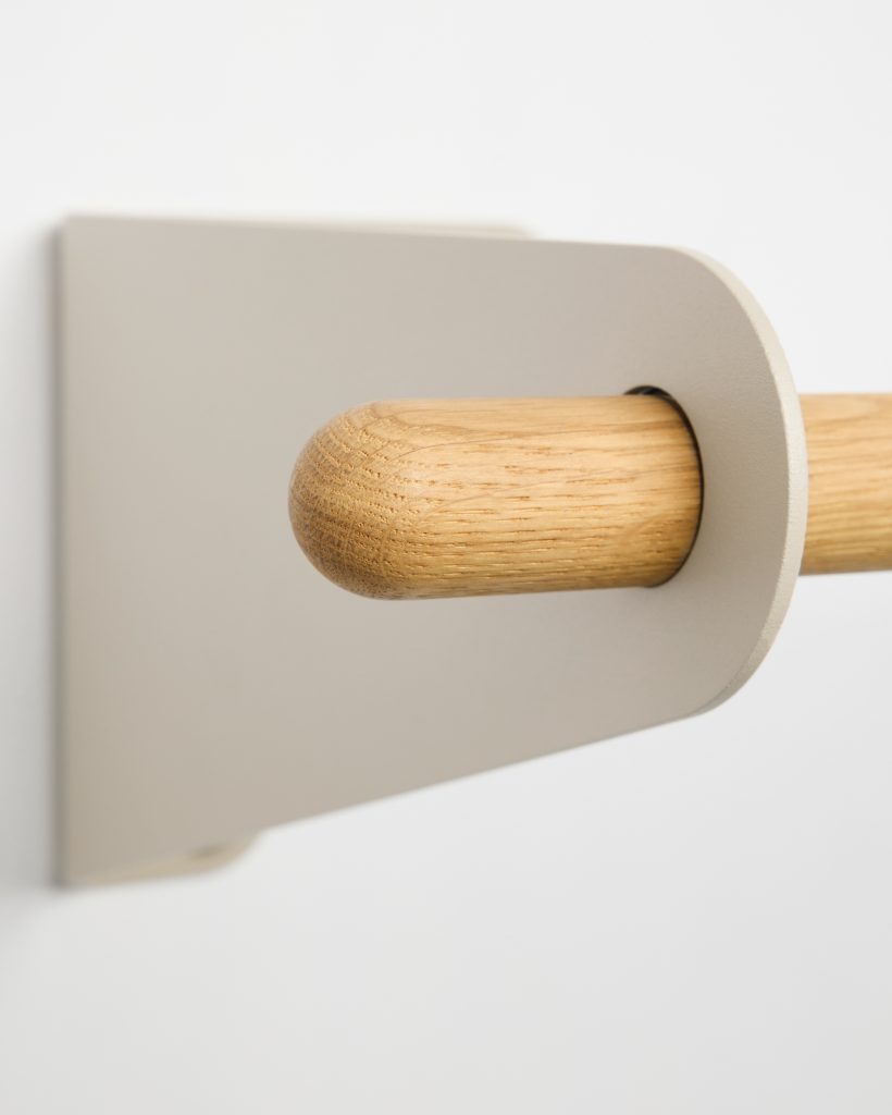 Close-up of a WoodUpp wall-mounted oak rail and matte beige metal bracket: a rounded oak dowel with visible grain slides through a rounded rectangular wall plate, showing smooth sanded wood end and minimalist powder-coated bracket detail against a white background.
