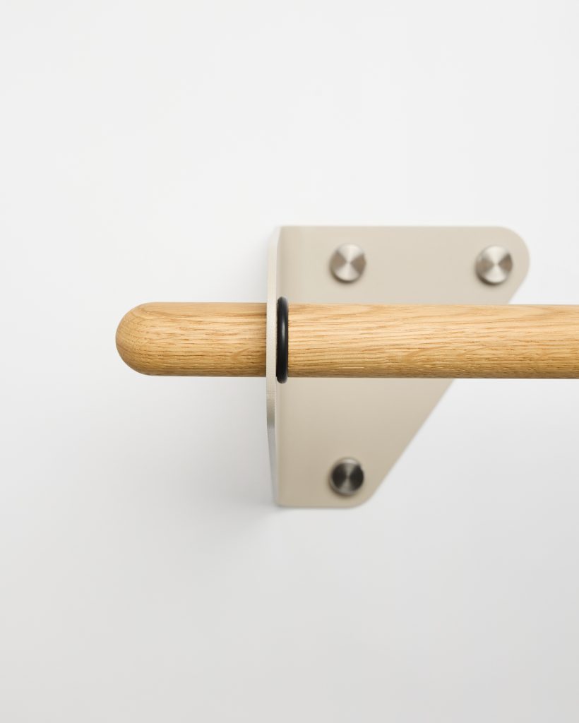 Close-up of a WoodUpp oak rail and matching wall bracket: a smooth rounded light-oak dowel inserted through a beige metal triangular bracket secured by three stainless bolts, with a small black rubber spacer ring between rod and bracket, mounted on a white wall.