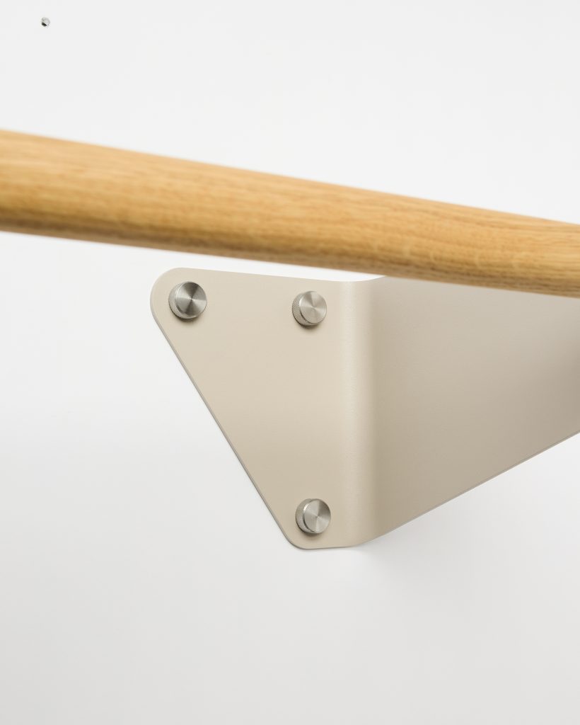WoodUpp wall-mounted handrail: close-up of a smooth oak handrail fitted into a beige, triangular powder-coated steel bracket from WoodUpp, secured by three brushed stainless steel cap screws. Minimal, modern detail shot showing the join of natural wood and matte metal hardware on a white wall.
