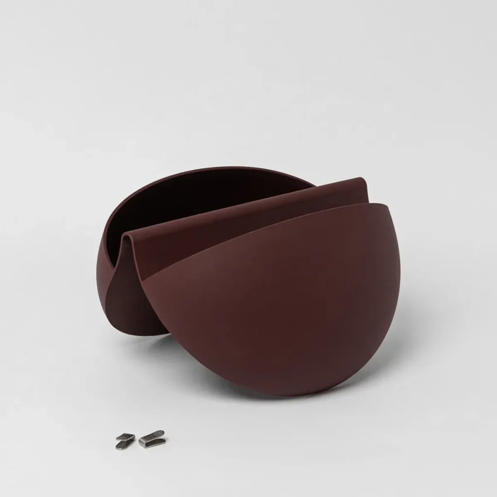 WoodUpp dark brown matte double-shell organizer sits on a white background, its two overlapping semi-spherical panels forming a sculptural storage bowl with a central slot. Three small brushed-metal clips sit nearby. Image showcases WoodUpp’s minimalist container and matching metal accessories.