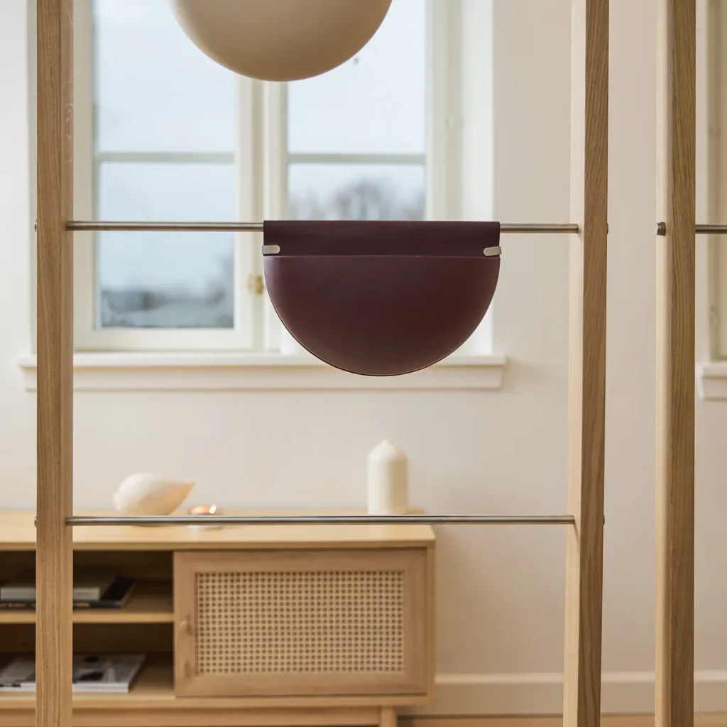 Minimal interior scene showing a WoodUpp oak frame with stainless-steel horizontal bars, featuring a maroon leather half‑moon hanging pouch attached with metal clips — a WoodUpp accessory — above a wooden sideboard with candle and decorative shell visible through a bright window.