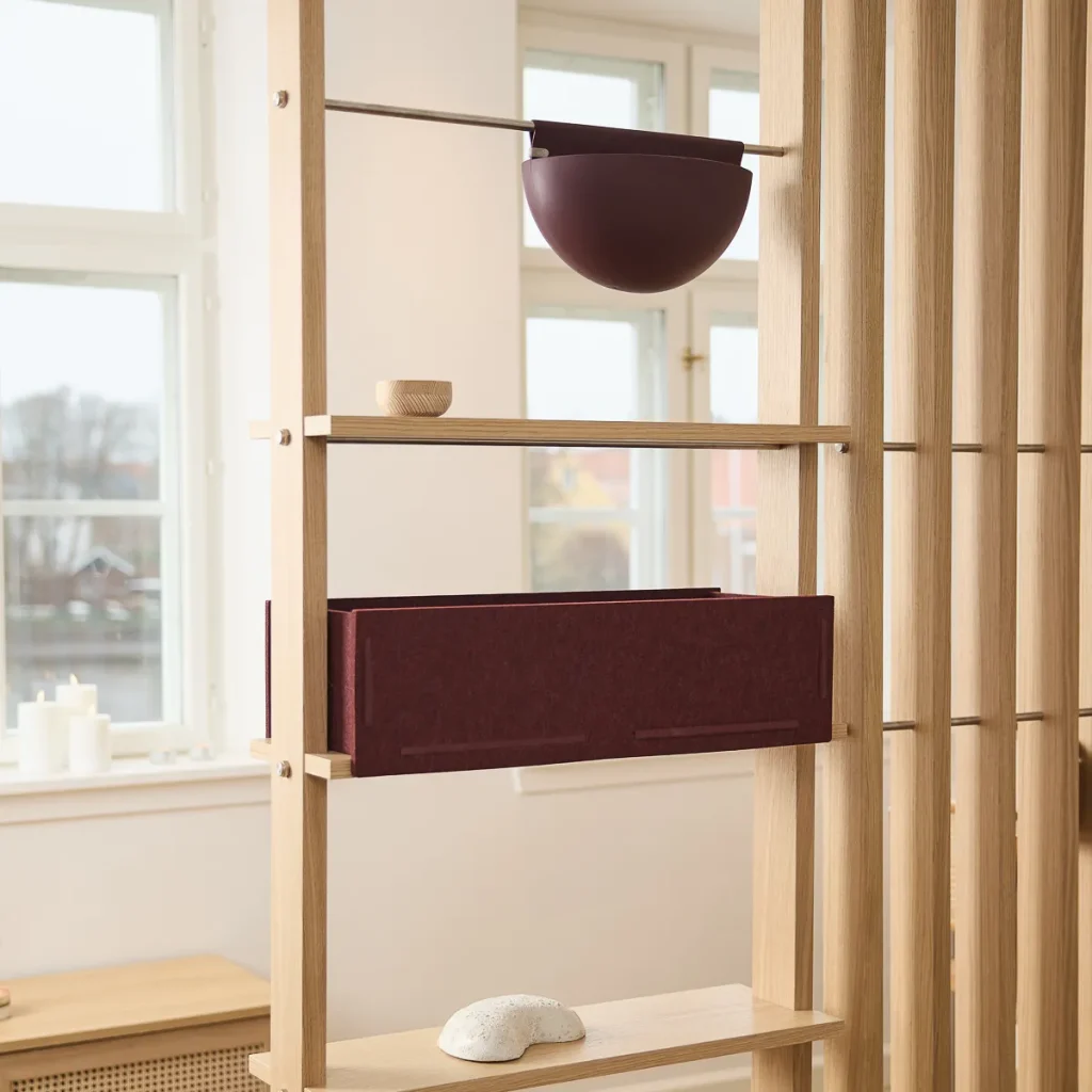 WoodUpp oak room-divider shelving unit with vertical slats and horizontal shelves displayed in a bright room. WoodUpp accessories include a burgundy felt storage box fitted on a shelf, a matching hanging half‑sphere planter suspended from a metal rod, plus a small wooden bowl and white pebble ornament.