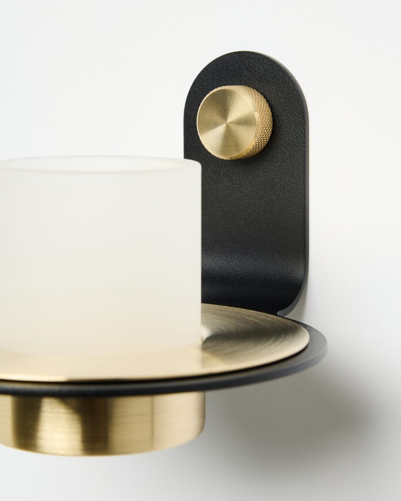 Close-up of a WoodUpp wall sconce mounted on a white wall: matte black curved metal bracket with a knurled brass adjustment knob, brushed brass circular plate and cup, and a satin frosted cylindrical glass shade. Clean, minimalist modern finishes and subtle textures are visible.
