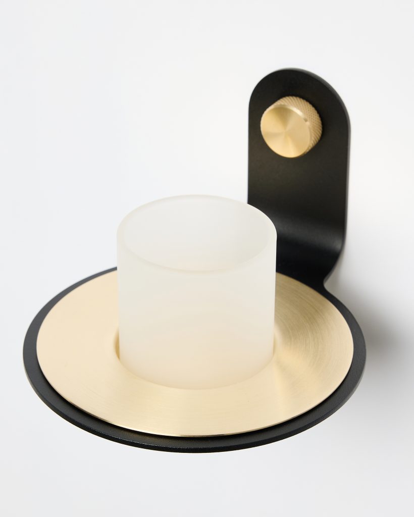 WoodUpp wall sconce featuring a black metal bracket with a knurled brass-tone adjustment knob, a round brushed brass-tone plate, and a frosted cylindrical glass shade resting in the center. Minimalist, modern finish showcasing contrast between matte black, warm brass, and translucent glass.