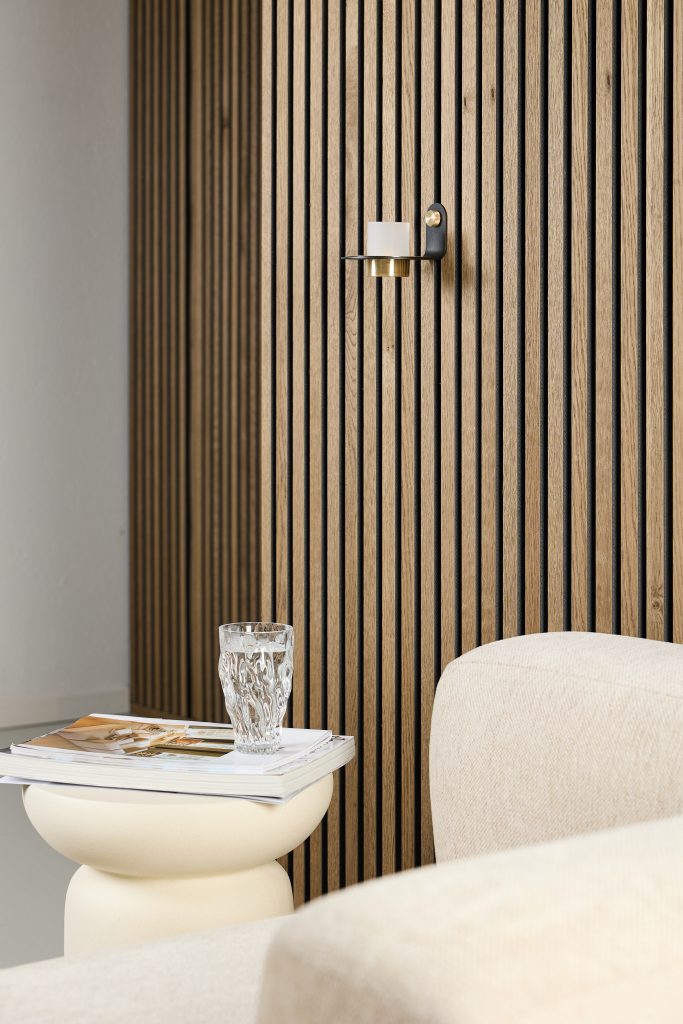 WoodUpp oak slat wall panels create a vertical, natural oak backdrop with narrow black shadow gaps. A WoodUpp black-and-brass wall candle sconce with a white pillar candle is mounted centrally. In foreground, a cream sculpted side table holds magazines and a textured water glass beside a beige sofa.