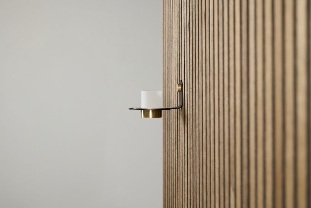 WoodUpp wall-mounted candle holder in black metal with a brass cup and frosted glass votive, attached to a WoodUpp oak vertical slatted wall panel, photographed against a soft neutral background, showcasing the brand's minimalist, modern hardware and timber slat design.