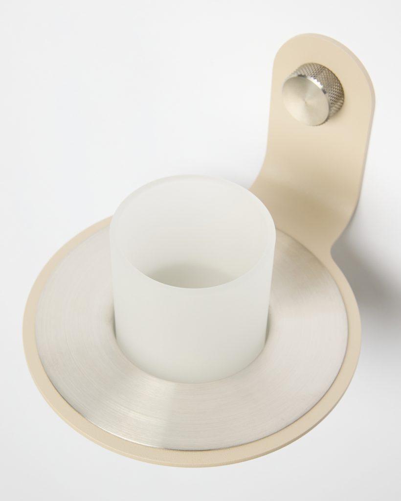 WoodUpp wall-mounted tumbler: close-up of a frosted glass cup seated in a circular brushed stainless-steel ring, set into a beige metal wall bracket with a textured metal mounting knob. Minimal, modern bathroom accessory showing material details and smooth matte finish.