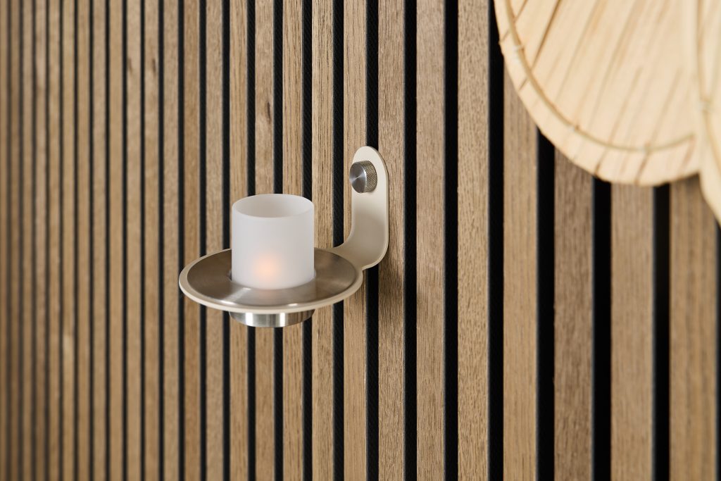 WoodUpp slatted wall panels in natural oak with black recessed grooves frame a minimalist steel wall-mounted candle holder from WoodUpp, featuring a brushed-metal plate and frosted glass LED tealight; the warm wood grain contrasts with the cool metallic sconce, creating a modern, cozy accent.