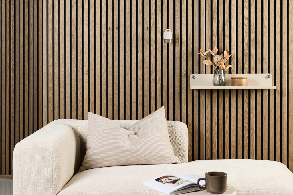 WoodUpp: a cozy seating corner featuring WoodUpp vertical oak slat wall panels with dark grooves, a mounted WoodUpp minimalist cream floating shelf holding a glass vase with dried leaves and a small wooden box, plus a beige sofa with cushion, open magazine and brown ceramic mug.